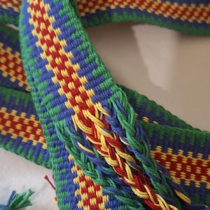Puede incluir: Un cinturón textil tejido en tonos verdes, azules, rojos y amarillos. El cinturón presenta un patrón geométrico y una sección trenzada con borlas. El diseño es intrincado y colorido, mostrando una artesanía tradicional.