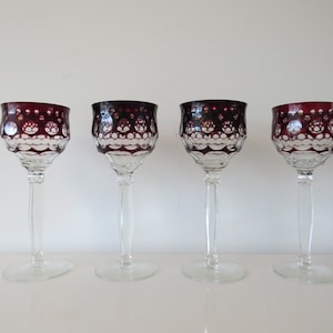 Op de afbeelding: Vier heldere wijnglazen met robijnrode kelken en heldere stelen. De kelken hebben een decoratief patroon van cirkels en sint-jakobsschelpen. De glazen worden gepresenteerd op een wit oppervlak tegen een witte achtergrond.