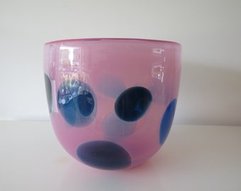 May include: A pink glass bowl with a rounded shape. The bowl is decorated with large, irregularly shaped blue and dark blue polka dots. The bowl is a decorative item.