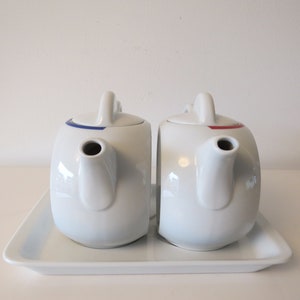 Op de afbeelding: Twee witte keramische theepotten met blauwe en rode accenten op een witte dienblad. De theepotten hebben een afgeronde vorm en een tuit aan de zijkant.