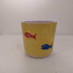 Puede incluir: Una taza de cerámica amarilla con un diseño de peces rojo y azul.