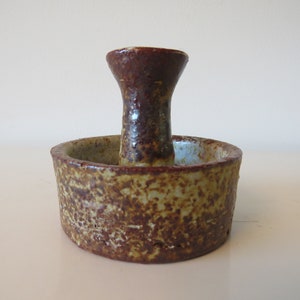 Puede incluir: Un portavelas de cerámica marrón y verde con un centro elevado para una vela. El portavelas tiene una superficie rugosa y texturizada.