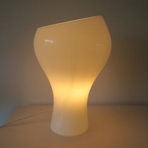 Könnte beinhalten: Eine weiße, moderne Tischlampe mit einem einzigartigen, geschwungenen Design. Die Lampe hat eine breite, abgerundete Oberseite, die sich zu einem schmaleren Sockel verjüngt. Die Lampe wird von innen beleuchtet und wirft ein warmes Licht. Die Lampe ist etwa 30 cm hoch.