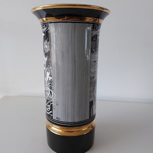 May include: Black and white ceramic vase with gold trim. The vase has a cylindrical shape and features a detailed design of stylized waves and floral motifs.