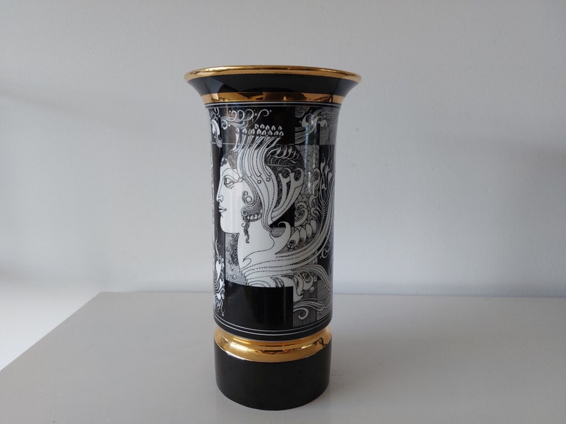 May include: Black and white ceramic vase with a gold rim and base. The vase features a detailed illustration of a woman's profile with intricate patterns and designs.