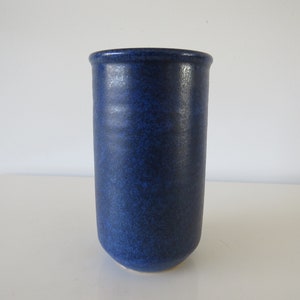 Könnte beinhalten: Eine hohe, zylindrische Keramikvase mit einer tiefblauen Glasur. Die Vase hat einen leicht abgerundeten Boden und eine glatte, gleichmäßige Oberfläche.