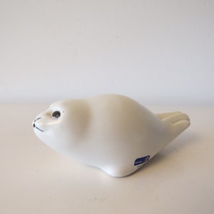 May include: White ceramic seal figurine with black eyes. The figurine is in a curled up position with its flippers tucked under its body. The figurine has a blue label on its side that reads "Rorstrand" and "Made in Sweden".