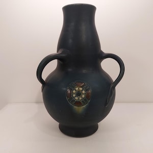 Puede incluir: Un jarrón de cerámica azul oscuro con dos asas y un diseño decorativo en el centro. El diseño presenta un círculo con formas geométricas en tonos de verde, marrón y blanco.