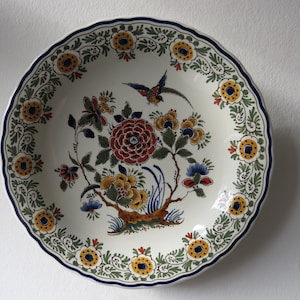 Può includere: Un piatto in ceramica bianca con un motivo floreale blu, verde, giallo e rosso. Il piatto presenta un uccello e un albero con fiori.