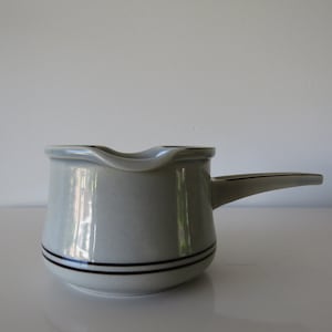 Op de afbeelding: Een grijze gravy boat van keramiek met een bruine streep rond de rand. De boot heeft een handvat en een tuit.