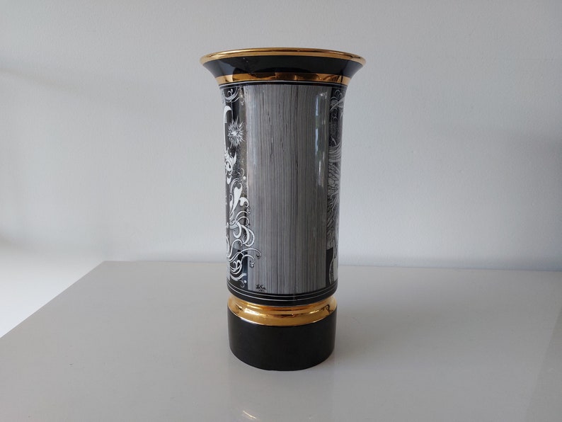 May include: Black and white ceramic vase with gold trim and a floral design. The vase has a cylindrical shape and a wide mouth.
