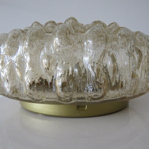 Op de afbeelding: Een heldere glazen plafondlamp met een getextureerd, geblazen oppervlak. De lamp heeft een gouden metalen basis.