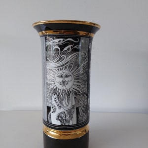 May include: A black and white ceramic vase with a gold rim and base. The vase features a stylized sun with a face and a swirling design.