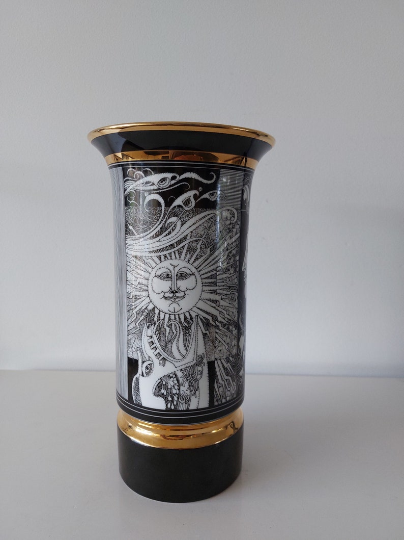 May include: A black and white ceramic vase with a gold rim and base. The vase features a stylized sun with a face and a swirling design.