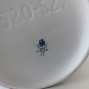May include: Close-up of the bottom of a white ceramic item with a blue and white logo that reads "Holl&oacute;h&aacute;za Hungary 1831 27" and the number "520-52".