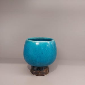 May include: A teal blue ceramic cup with a wide base. The cup has a smooth, glossy finish and a rounded shape.