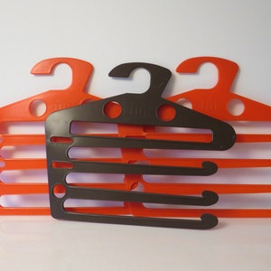 Op de afbeelding: Set van drie plastic hangers, twee zijn oranje en één is bruin. De bruine hanger heeft vijf gleuven om kleding op te hangen. De hangers zijn ontworpen om ruimte te besparen in een kast.