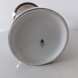 May include: The bottom of a white ceramic vase with a blue and gold design. The vase has a blue eagle logo with the text "Holl&oacute;h&aacute;za Hungary 1836 27" and the number "520-57".
