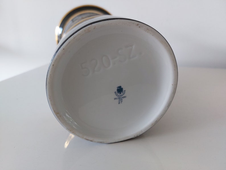 May include: The bottom of a white ceramic vase with a blue and gold design. The vase has a blue eagle logo with the text "Holl&oacute;h&aacute;za Hungary 1836 27" and the number "520-57".