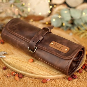 May include: A rolled brown leather tool roll with a buckle closure and a small leather tag with the initials "K.L." is displayed on a wooden plate. The tool roll is surrounded by tools and decorative elements.