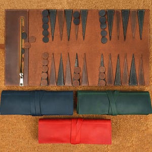 May include: A brown leather backgammon set with black and brown game pieces. The set is open, revealing the playing surface. Below are three rolled-up leather cases in navy blue, green, and red.