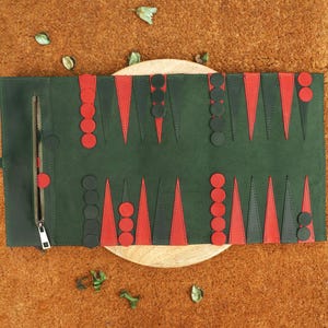 May include: A rolled-up backgammon board in dark green leather with red and black triangular points and circular game pieces. The board is unrolled on a wooden surface, with a zipper closure visible on the side.