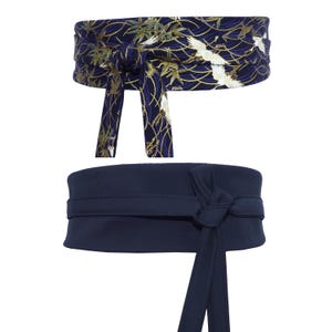 Könnte beinhalten: Zwei Obi-Gürtel. Der obere Gürtel ist marineblau mit einem goldenen und weißen Kranich- und Blättermuster. Der untere Gürtel ist einfarbig marineblau. Beide Gürtel sind mit einem Knoten und langen Bändern gebunden.