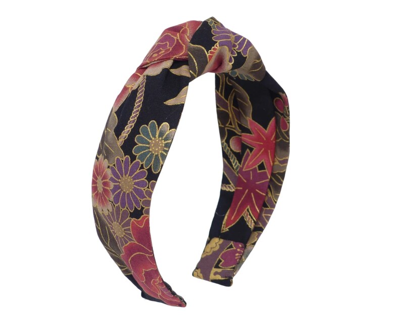 May include: A black headband with a knotted design. The headband is covered in a floral print with red, pink, gold, and purple flowers on a black background.