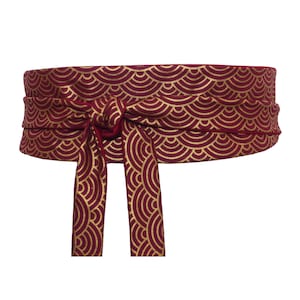 Può includere: Una cintura in tessuto rosso con un motivo geometrico dorato. La cintura è annodata in un fiocco sul davanti.