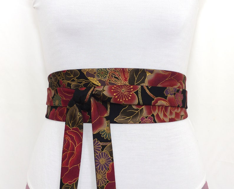 May include: A black fabric belt with a floral pattern in shades of red, gold, and purple. The belt is tied in a bow at the front.