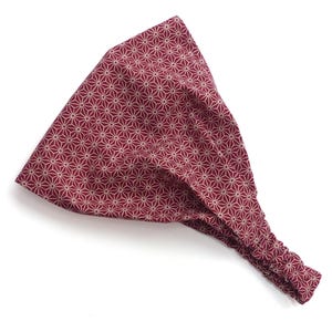 May include: A burgundy headband featuring a geometric starburst pattern in a beige hue. The back is elasticated for a comfortable fit. This hair accessory is suitable for various hairstyles and is approximately 20 cm wide.