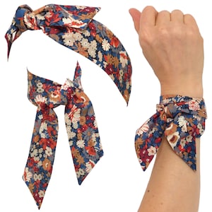 Könnte beinhalten: Set aus floralen Accessoires: Stirnband, Krawatte und Armband. Der Stoff zeigt ein Muster aus kleinen Blumen in Rot-, Blau-, Braun- und Cremetönen. Das Stirnband ist mit einer Schleife gebunden, die Krawatte hat lange Enden und das Armband ist um ein Handgelenk gebunden.