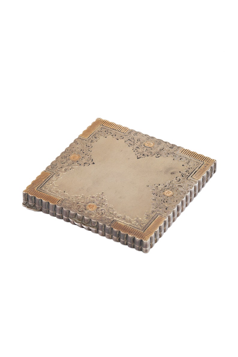 Puede incluir: Un estuche compacto de plata y oro con un dise&ntilde;o grabado ornamentado. El estuche tiene un borde festoneado y una tapa con bisagras.