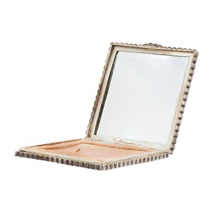 Puede incluir: Un espejo compacto de plata con un borde festoneado y un interior de terciopelo rosa. El espejo est&aacute; abierto y la tapa est&aacute; levantada.