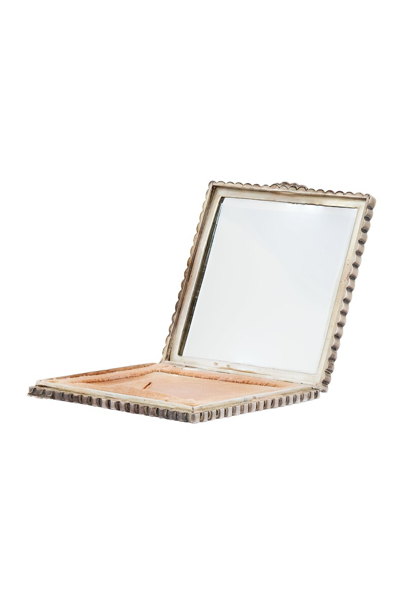 Puede incluir: Un espejo compacto de plata con un borde festoneado y un interior de terciopelo rosa. El espejo est&aacute; abierto y la tapa est&aacute; levantada.