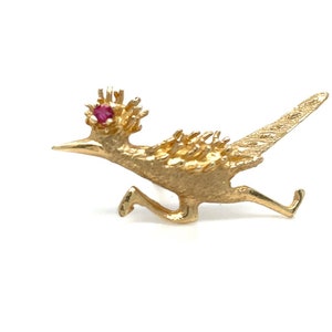 May include: A gold-toned roadrunner pin with a red gemstone eye. The roadrunner is depicted in a running pose with its wings spread out.