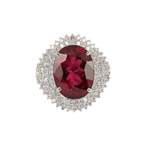May include: A ring with a large oval-shaped red gemstone surrounded by a halo of smaller white gemstones.