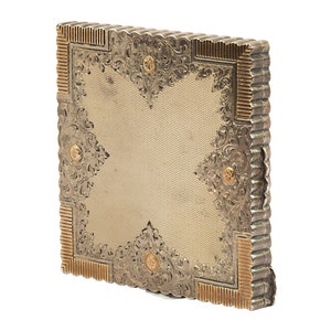 Puede incluir: Un estuche compacto de plata y oro con un dise&ntilde;o ornamentado. El estuche es de forma cuadrada y est&aacute; decorado con motivos florales y detalles dorados.