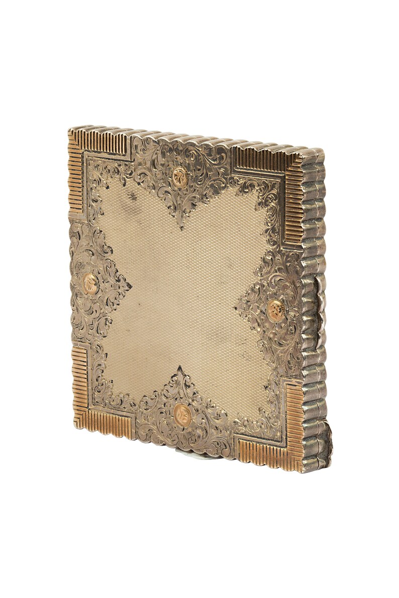 Puede incluir: Un estuche compacto de plata y oro con un dise&ntilde;o ornamentado. El estuche es de forma cuadrada y est&aacute; decorado con motivos florales y detalles dorados.