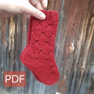Könnte beinhalten: Rote Stricksocken mit einem Spitzenmuster. Die Socken werden in einer Hand vor einem Holzgrund gehalten. Der Text "PDF" ist in der linken unteren Ecke des Bildes sichtbar.
