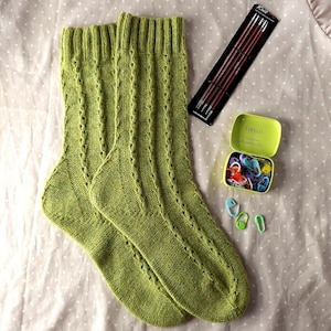 Könnte beinhalten: Ein Paar hellgrüne Stricksocken mit Rippenmuster. Stricknadeln und Maschenmarkierer liegen neben den Socken. Eine kleine grüne Dose trägt die Aufschrift "Compass" und "You know an apple a day..."