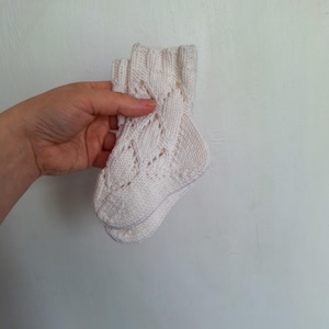 以下が含まれることがあります： 繊細なケーブル編みのパターンが施された、白いニットのベビーソックスのペアです。