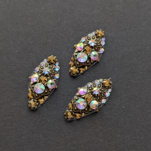 May include: Three decorative, oval embellishments. Each has a bronze-coloured metal base, decorated with iridescent, round and star-shaped rhinestones and smaller, amber-coloured stones. They are arranged on a dark grey surface.