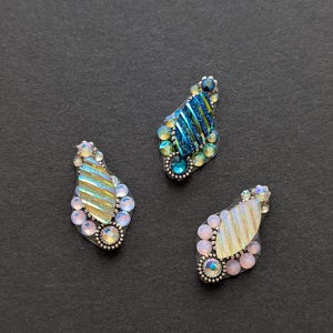 May include: Three decorative, teardrop-shaped embellishments with iridescent, striped centers. Each piece is adorned with colorful rhinestones and a silver-toned, beaded border. The embellishments are arranged on a dark gray surface.