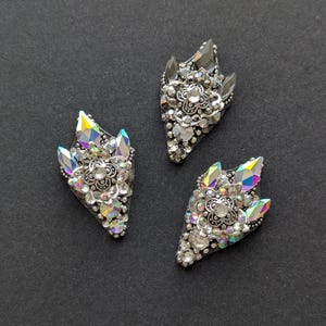 May include: Three ornate, silver-toned embellishments with iridescent crystal accents. Each piece features a central floral design surrounded by sparkling rhinestones and pointed crystal shapes, set against a dark background.