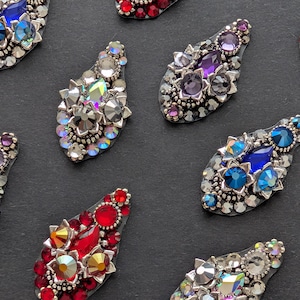 May include: Close-up of several decorative nail art pieces. Each piece is a teardrop shape, encrusted with various colourful rhinestones and crystals in shades of red, blue, purple, and clear. The pieces are set against a dark background.