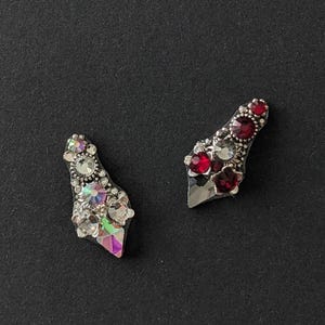 May include: Two silver-toned, jeweled, decorative embellishments. One is adorned with clear iridescent rhinestones, the other with red rhinestones.