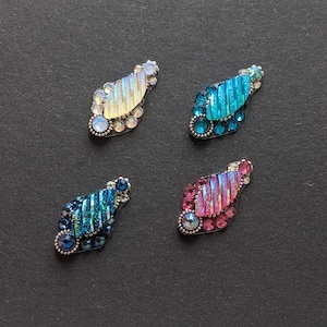 May include: Four decorative, jewel-encrusted embellishments in a teardrop shape. Each has a central, textured section in shades of blue, pink, and yellow, surrounded by small, clear and coloured rhinestones. The embellishments are set against a dark grey background.