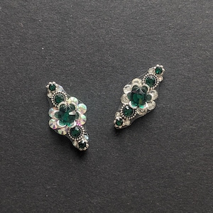 May include: Two decorative, silver-toned embellishments with a floral design. Each features a central emerald-green gemstone surrounded by clear, iridescent crystals and smaller green stones, set against a dark background.