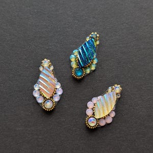 May include: Three decorative embellishments in a teardrop shape, each with iridescent and jewel-toned details. They feature a central striped design, surrounded by small gems and a gold-coloured border. The background is a dark, solid colour.